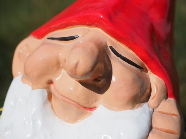 A close-up of a ceramic garden gnome with a red hat, white beard, closed eyes, and a slight smile, set against a blurred green background.