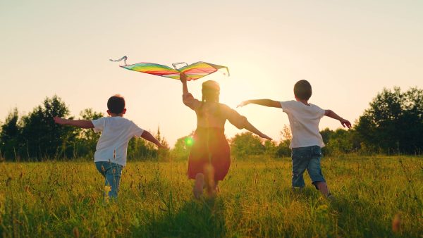 Three children run through a grassy field at sunset, with one holding a colorful kite above their head. The sun is low in the sky, creating a warm, golden glow over the scene.