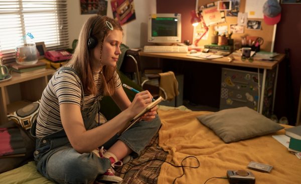 A teenage girl sits cross-legged on her bed, wearing headphones and writing in a notebook. Her room is decorated with retro items, including a desktop computer, posters, and string lights. Sunlight streams through the blinds.