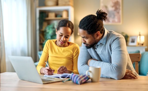 A young girl in a yellow shirt writes in a notebook while an adult man sits beside her, watching and smiling. They are at a table with a laptop, mug, and pencil case in a cozy room with shelves and soft lighting.