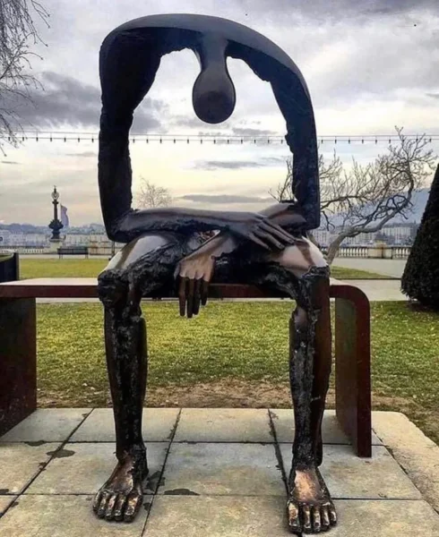 A sculpture of a seated, headless figure with slumped shoulders and arms crossed over its knees, made of dark metal, displayed outdoors on a stone platform with grass and trees in the background.