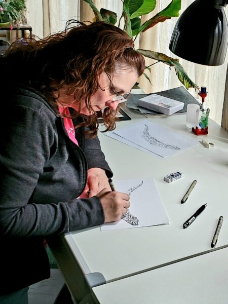 A woman with wavy brown hair and glasses draws intricate designs on paper at a white desk, surrounded by markers, an eraser, and a desk lamp, with houseplants in the background.