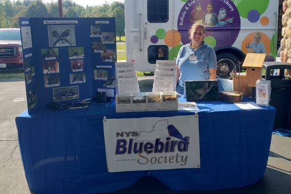 A. T. Baron behind a table with a NYS Bluebird Society banner, brochures, bird information displays, and birdhouse models. An educational van and trees are visible in the background.